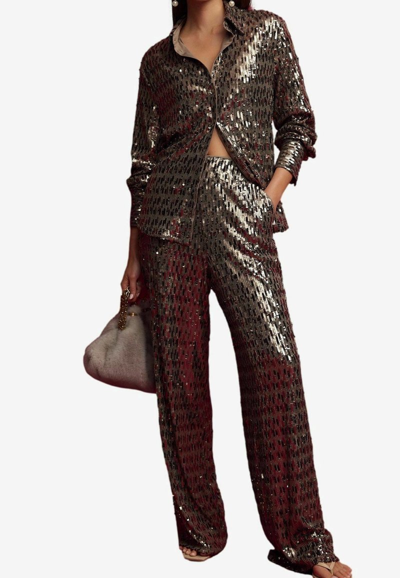 Femme portant une chemise à manches longues en sequins assortie et un pantalon à jambes larges, tenant un sac à main duveteux, debout avec une main dans la poche.