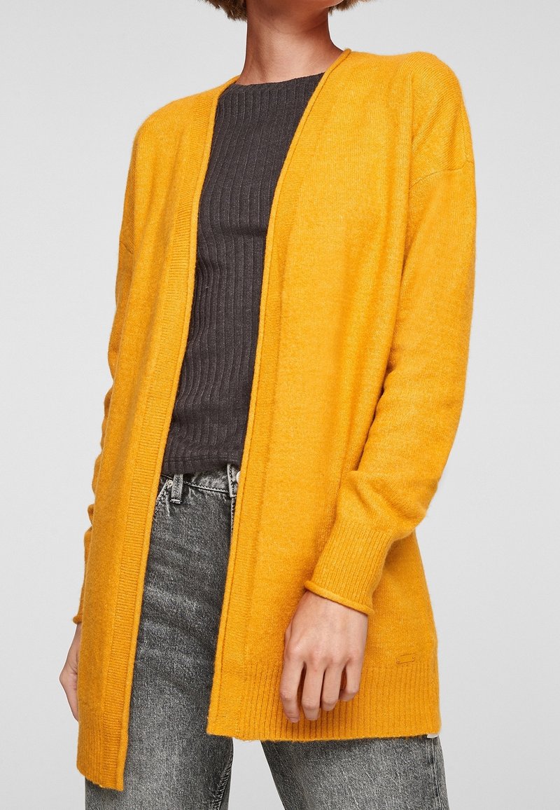Femme portant un cardigan moutarde ouvert sur un t-shirt gris foncé côtelé et un jean noir délavé, sur un fond uni.