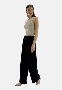 Beżowy, krótki top na ramiączkach i czarne szerokie spodnie, wykonane z miękkiego materiału; modelka nosi buty w kolorze cielistym; minimalistyczny design.