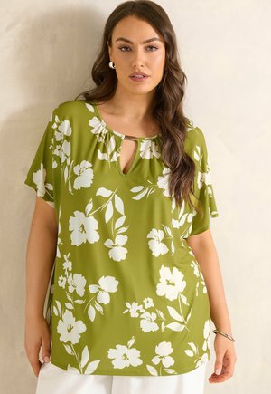 Vrouw met lang bruin haar die een groene blouse met wit bloemenpatroon en korte mouwen draagt, staand tegen een lichte muur.