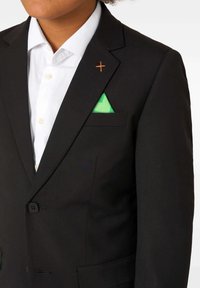 Cilvēks, kuram mugurā ir melns uzvalka žakets ar oranžu izšūtu krustu pie atloka, balta pogājama krekla un zaļa kabatas laukuma.