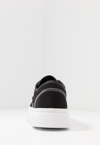 Černé plátěné tenisky s bílým dvojitým švem a silnou bílou podrážkou s vyraženým textem, zobrazené zezadu na bílém pozadí.