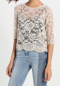 Blusa in pizzo con motivo floreale, maniche a tre quarti, scollatura tonda e orlo a onda. Il colore è beige chiaro. Indossata con jeans blu.