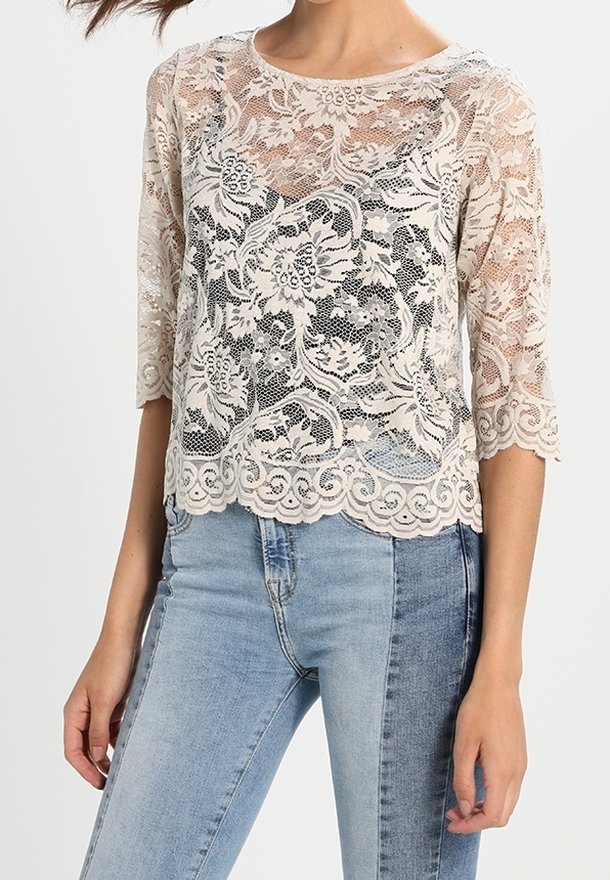 Blusa in pizzo con motivo floreale, maniche a tre quarti, scollatura tonda e orlo a onda. Il colore è beige chiaro. Indossata con jeans blu.