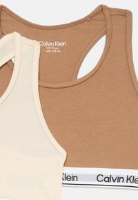 Du trijų medvilnės mišinio liemenėlės: viena šviesiai bežinė, kita ruda, su juodu ir baltu Calvin Klein logotipo juosmeniu, apvalaus kaklo dizainas.