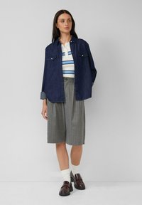 Donna in piedi con le mani in tasca, che indossa una giacca di jeans scura, una maglietta a righe bianche e blu, pantaloni larghi grigi a righe, calzini bianchi e mocassini scuri.