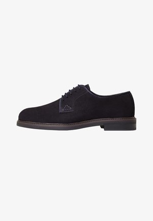 Chaussure oxford en suede noir avec un bout rond, un laçage, des coutures contrastantes et une semelle en caoutchouc plate. Un style facile à porter au quotidien.