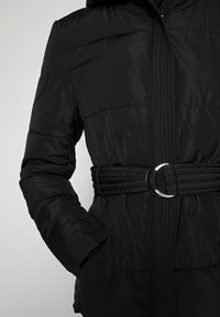 Sort puffet jakke med hætte, der har en indsnøret talje med en sølvring, quiltet tekstur og lynlås foran.