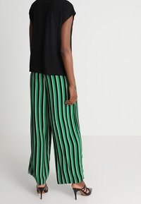 Svart kortärmad topp kombinerad med vida byxor i gröna och svarta ränder. Byxorna har vertikala ränder och en flödande silhuett. Svarta klackar.