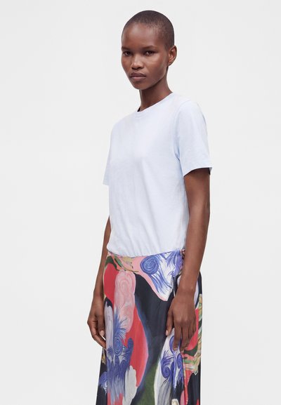 Femme aux cheveux courts portant un t-shirt blanc uni et une jupe colorée avec des motifs floraux abstraits, debout devant un fond clair uni.