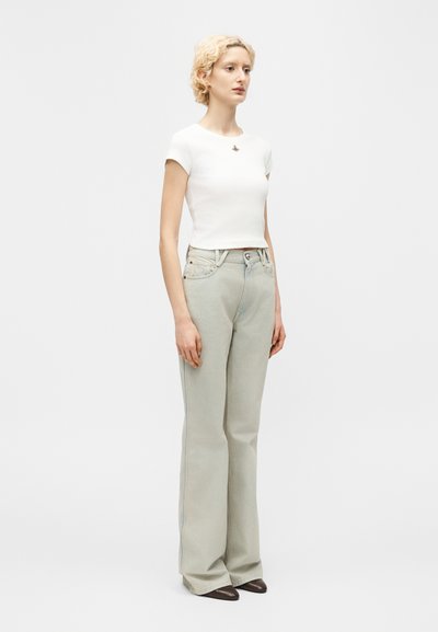 Γυναίκα με κοντά σγουρά ξανθά μαλλιά φοράει λευκή εφαρμοστή κοντομάνικη μπλούζα, ανοιχτό γκρι παντελόνι με ψηλή μέση και φαρδιά μπατζάκια, καθώς και σκούρα μυτερά παπούτσια.