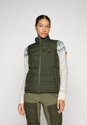 Grønn puffer vest med glidelås foran, høy krage og lommer; brukt over en mønstrete genser og kombinert med olivengrønne bukser.