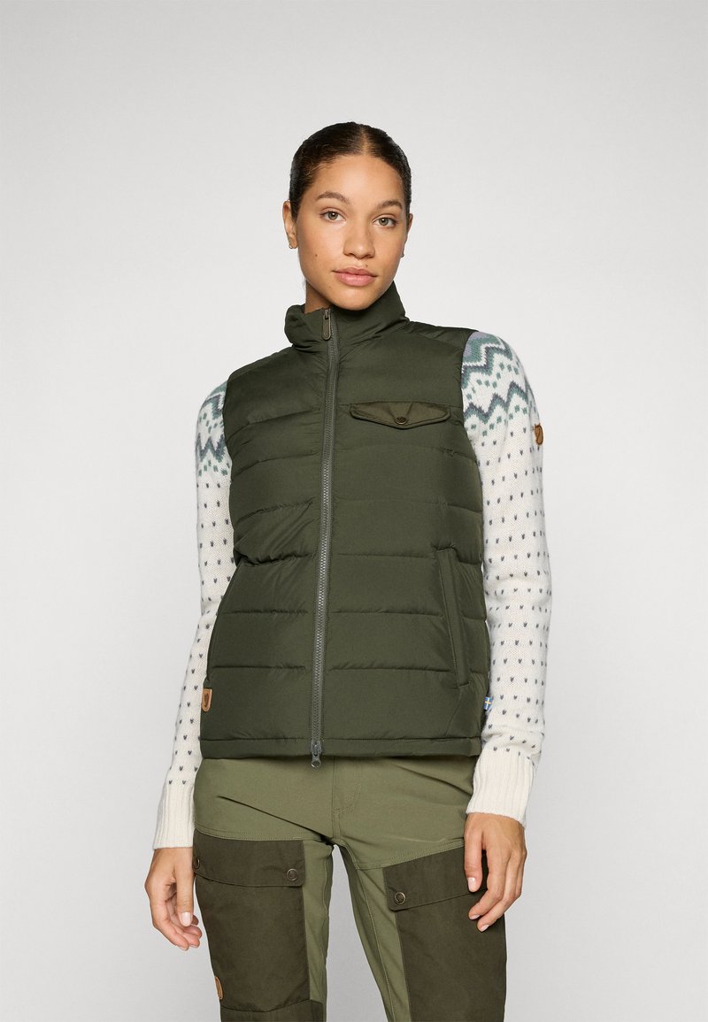 Groene puffer vest met een ritssluiting aan de voorkant, hoge kraag en zak; gedragen over een gebreid trui en gecombineerd met olijfgroene broek.