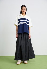 Kvinna som står på grön golv och bär en vit och marinblå skjorta, lång svart kjol och beige sneakers med ljusblå ränder.