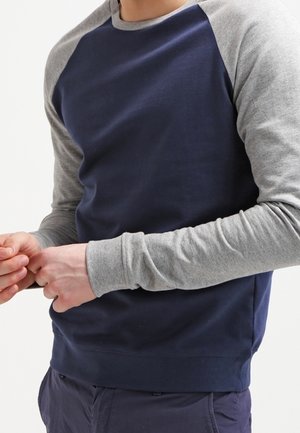Homme portant une chemise bleu marine avec des manches raglan gris clair, tenant ses mains jointes devant un fond blanc.