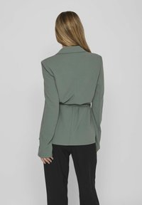 Grön kavaj med långa ärmar och skräddarsydd passform, med bälte vid midjan, slät yta och vassa slag, bärs över svarta byxor.