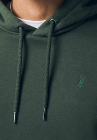 Gros plan d'une personne portant un sweat à capuche vert foncé avec des cordons et un petit logo de cerf brodé sur la poitrine, une chemise blanche visible en dessous.