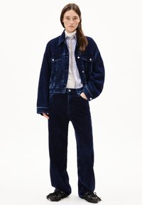 Kamelångerjacka och byxor i mörk marinblå med vertikal ribbtextur. Vit skjorta med subtila vertikala ränder. Svarta fransade skor.