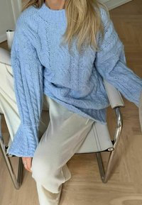 Person i ljusblå tjockstickad tröja och vita byxor sitter på en vit läder- och kromstol på ett trägolv.