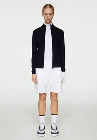 Ναυτική ζακέτα με φερμουάρ, λευκή μπλούζα από τζέρσεϊ, λευκά σορτς και γάντια. Το μοντέλο φορά ναυτικά και λευκά sneakers και λευκές κάλτσες μέχρι τον αστράγαλο με μαύρες ρίγες.