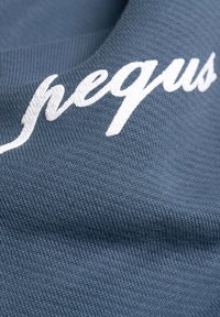 Svetlomodrá tkanina s textúrovaným vzorom, na ktorej je biela kurzíva s vyšívaným nápisom „pequs“. Detailný pohľad na materiál a dizajnové detaily.