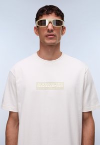 Απαλό μπεζ βαμβακερό t-shirt με κοντά μανίκια, που διαθέτει ένα υφασμένο ορθογώνιο λογότυπο σε αντίθετο χρώμα στο ύψος του στήθους.