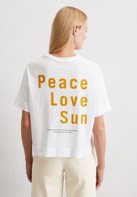 Weißes Baumwoll-T-Shirt mit einem verkürzten Design. Auf der Rückseite befindet sich ein auffälliger gelber Schriftzug "Peace Love Sun", ergänzt durch zusätzlichen Text darunter.