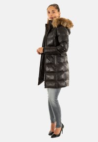 Abrigo acolchado negro con capucha de piel sintética, bolsillos con cremallera y un diseño acolchado, combinado con jeans claros y tacones negros.