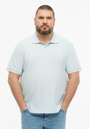 Homme avec barbe et cheveux attachés en arrière, portant un polo bleu clair et un jean, debout les mains dans les poches devant un fond blanc uni.
