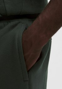 Tmavozelená látková vrecko na odeve s viditeľnou rukou, ktorá odpočíva vo vnútri. Mäkka textúra s prešitými okrajmi, ukazujúcí neformálny dizajn.