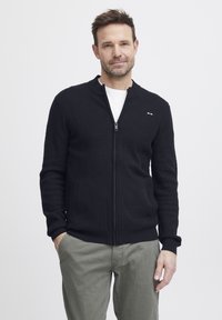 Svart glidelås cardigan med et teksturert strikkemønster, rund krage og ribbestrikkede mansjetter. Brukt over en hvit skjorte og stylet med lysegrå bukser.