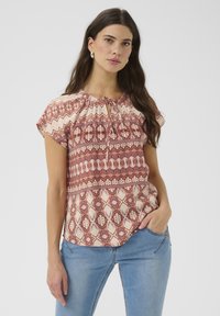 Kurzärmeliges, leichtes Oberteil in Creme und Rost mit geometrischen Mustern, gerafftem Ausschnitt und Schlüssellochdetail vorne. Kombiniert mit blauen Jeans.