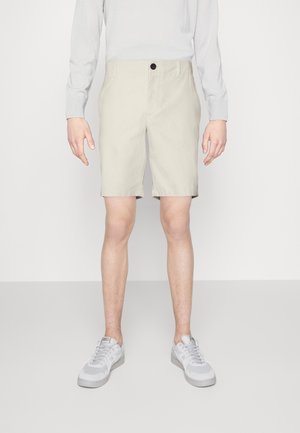 Homme portant un short beige au niveau des genoux, une chemise blanche à manches longues et des baskets grises, debout devant un fond uni.