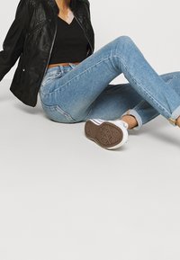 Chaqueta de piel sintética negra, top negro de cuello en V, jeans ajustados de mezclilla azul claro, zapatillas blancas con suelas marrones, puños enrollados y detalles mínimos.