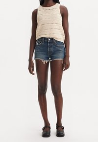 Canotta beige lavorata a maglia con strisce orizzontali e dettagli ritagliati, abbinata a pantaloncini di denim blu con orlo sfrangiato e finitura sbiadita.