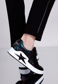 Stopa w czarnej sneakerze z białą podeszwą, ozdobionym czarną gwiazdą i błyszczącą dekoracją w postaci pszczoły na pięcie, w zestawieniu z czarnymi spodniami z paskiem z rypsowego kamienia.