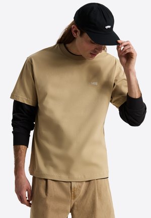 Mand iført beige Vans T-shirt lag-på-lag over en sort langærmet skjorte, beige bukser og en sort kasket, mens han justerer kasketkanten.
