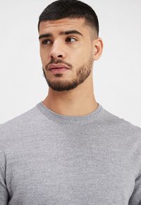 Grå stickad tröja med rund halsringning, mjuk textur och minimalistisk design. Har en liten logotyp på bröstet.