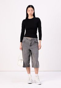 Schwarzes Langarmshirt, graue Cropped-Jeans und weiße High-Top-Sneaker. Das Model trägt eine kleine weiße Handtasche. Einfaches, lässiges Outfit.