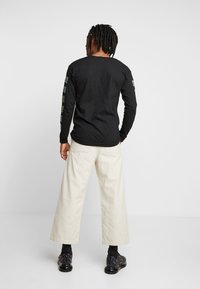 Černé tričko s dlouhým rukávem a zelenými grafickými detaily na rukávech, spárované s krémovými kalhotami s širokými nohavicemi a černými teniskami.