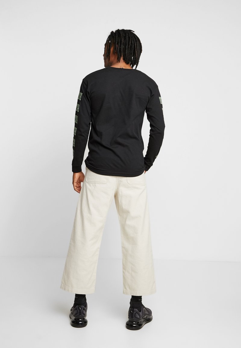 Černé tričko s dlouhým rukávem a zelenými grafickými detaily na rukávech, spárované s krémovými kalhotami s širokými nohavicemi a černými teniskami.