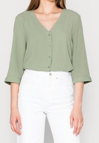 Lys grøn, V-hals bluse med trekvart ærmer og fem knapper ned ad forsiden, parret med hvide højtaljede jeans. Glat tekstur.
