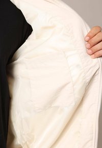 Veste légère de couleur crème à l'intérieur, avec une texture lisse et une poche plaquée sur le côté gauche, dotée d'une fermeture éclair.