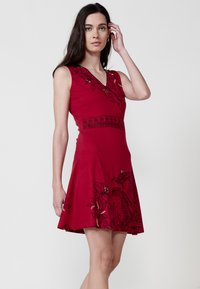 Rotes ärmelloses Kleid mit V-Ausschnitt, verziert mit schwarzen Blumenmustern und einem detaillierten Taillenbund; aus glattem Stoff mit einem weit schwingenden Rock.
