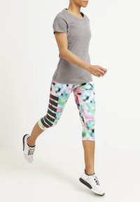 Osoba jde a má na sobě šedé tričko s krátkým rukávem, vícebarevné legíny po kolena se černými pruhy a bílé sportovní boty s černými podrážkami.