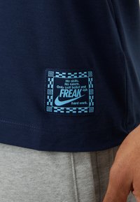 Marineblå bomulds T-shirt med et mønstret kantmærke, der har lyseblå typografi og Nike-logoet, med teksten "FREAK-ish hårdt arbejde."