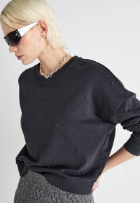 Svart genser med runde detaljer, rund hals og ribbestrikkede mansjetter. Sammen med et grått strikket skjørt og en sølvfarget kjede.