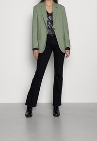 Blazer vert clair sur un haut à motif serpent noir, associé à un jean noir évasé et des bottines noires. Tissu lisse et design structuré.