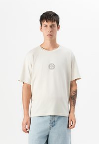 Man wearing a beige T-shirt with a small sun design in the center and light blue jeans, standing against a plain white background.