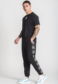 Svart t-shirt med en liten grafik på bröstet, tillsammans med svarta joggers med ett vitt mönster längs sidorna. Blå sneakers.